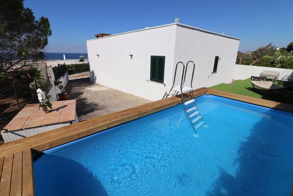 A swimming pool with a wooden deck, a white building with two doors, and a view of the sea.