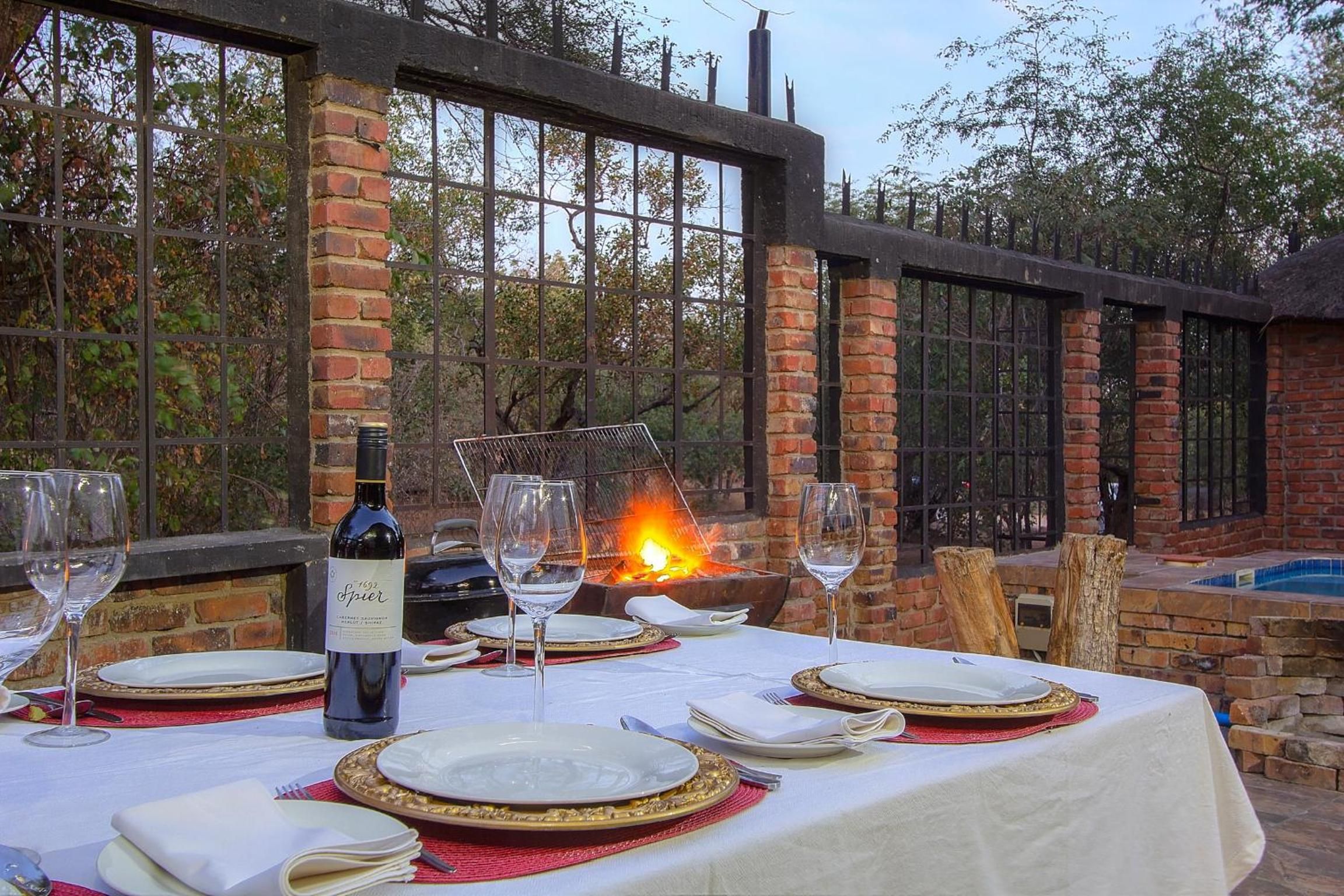 An outdoor dining area with a brick wall, a fire feature, and a pool in the background.
