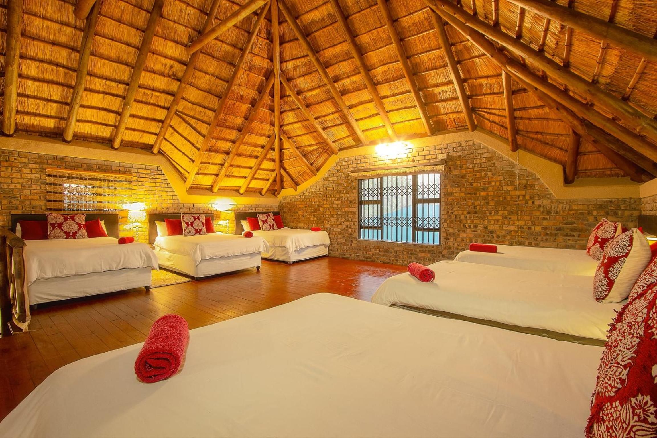 A hotel room with a high thatched ceiling, brick walls, and a large window with bars. There are four beds with white bedding and red pillows.