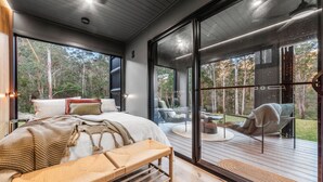 A modern bedroom with a large bed, a bench, and a view of a deck and forest. - Mijo | The Nook-Off grid Rural Retreat (Tenterfield)