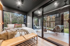 A modern bedroom with a large bed, a bench, and a view of a deck and forest.