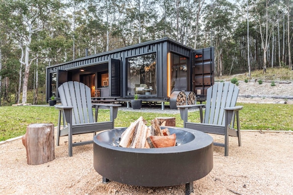 A modern cabin with a fire pit, two Adirondack chairs, and a dining table.