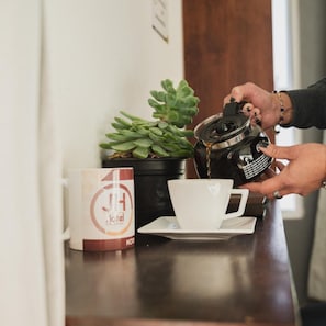 Una persona abocant cafè en una tassa al taulell, amb una planta en test i una tassa d'hotel.