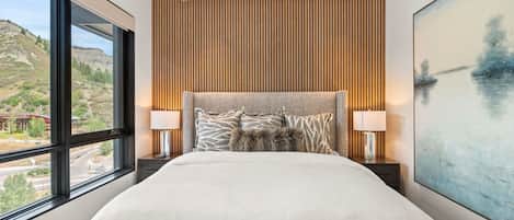 Un dormitorio moderno con una cama grande, dos mesitas de noche, vista a un paisaje a través de un ventanal amplio y una pared revestida de madera.