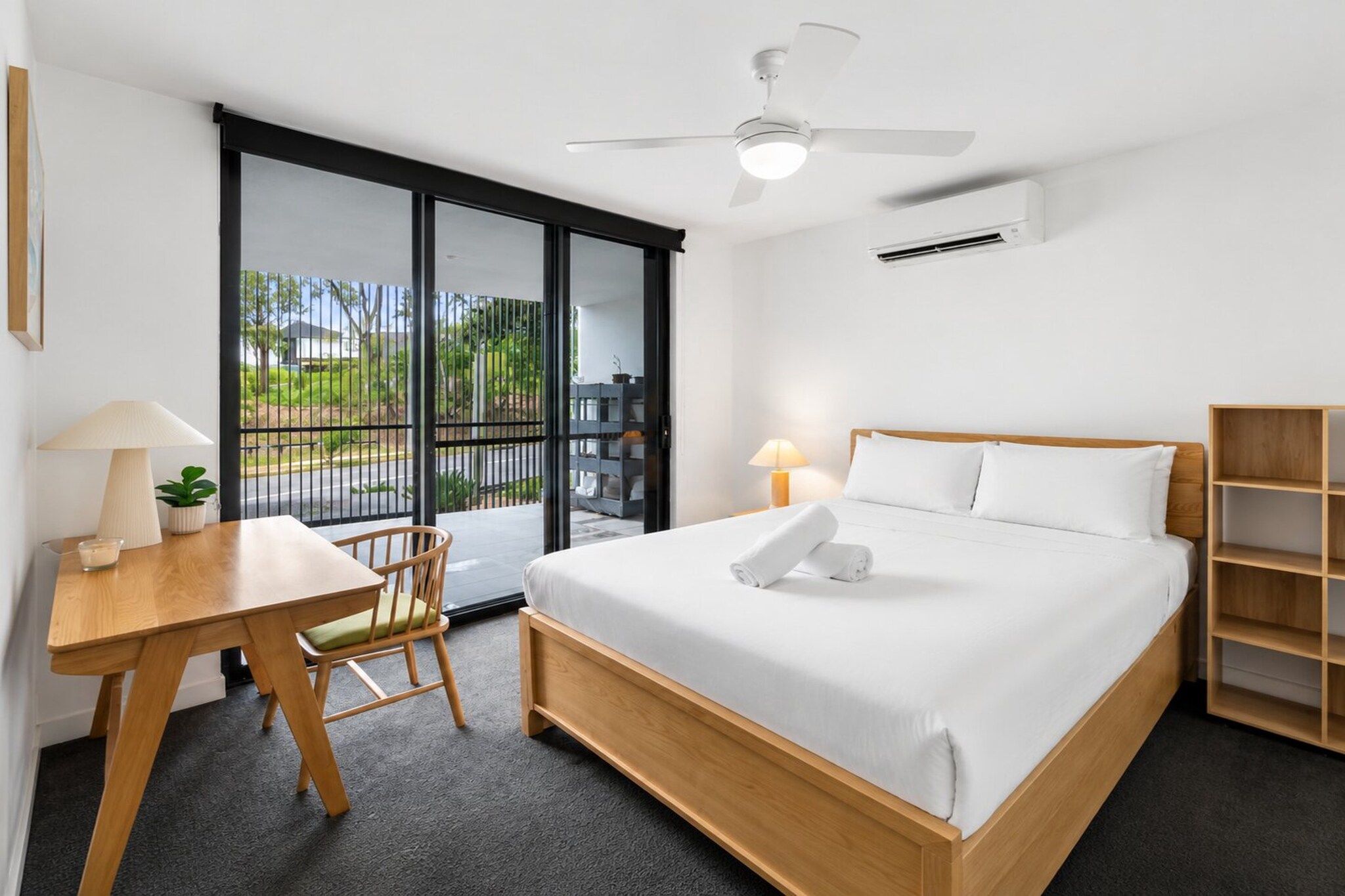 A hotel room with a bed, a desk, a chair, a ceiling fan, and a view of the outdoors.