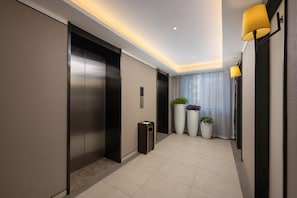 Un ascenseur moderne doté d’un panneau de commande, d’une poubelle et de plantes en pot, situé dans un couloir au sol de couleur claire et au plafond équipé de luminaires encastrés.