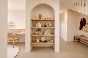 Un interior minimalista con una estantería central que exhibe diversos objetos decorativos, un banco de madera y un comedor con una silla y una mesa.