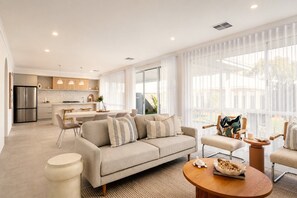 Een moderne woonkamer met een bank, stoelen en een salontafel. In de achtergrond een keuken met een koelkast en een eethoek.