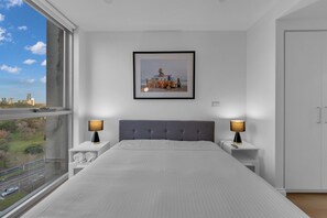 Quarto moderno com cama grande, duas mesas de cabeceira com abajures, um quadro na parede e vista da cidade através de uma janela ampla.