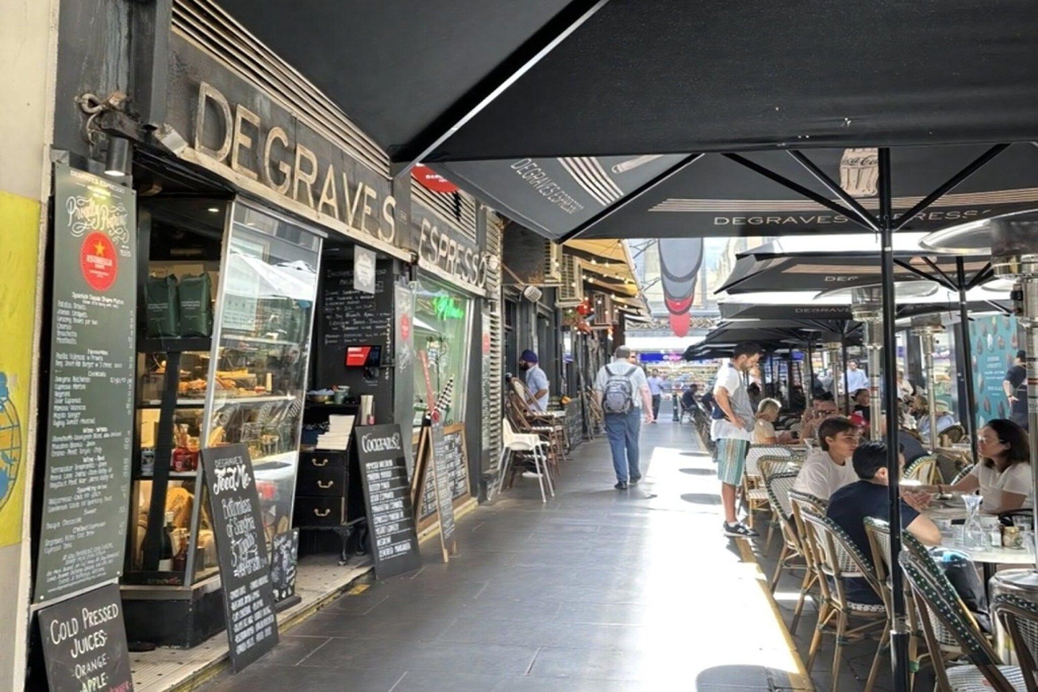 Um café ao ar livre com mesas e cadeiras, um letreiro com 'DEGRAVES' espresso e um quadro de menu.