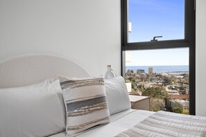 Ein Bett mit weißer Bettwäsche und gestreifter Kopfkissen, Blick auf eine Stadtkulisse durch ein Fenster und eine weiße Wand.