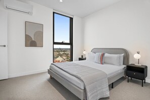 A bedroom with a large bed, a nightstand, a wall-mounted air conditioner, a window with a view, and a framed artwork. - Balcony 3-Bed Apartment Near Albert Park (St Kilda)