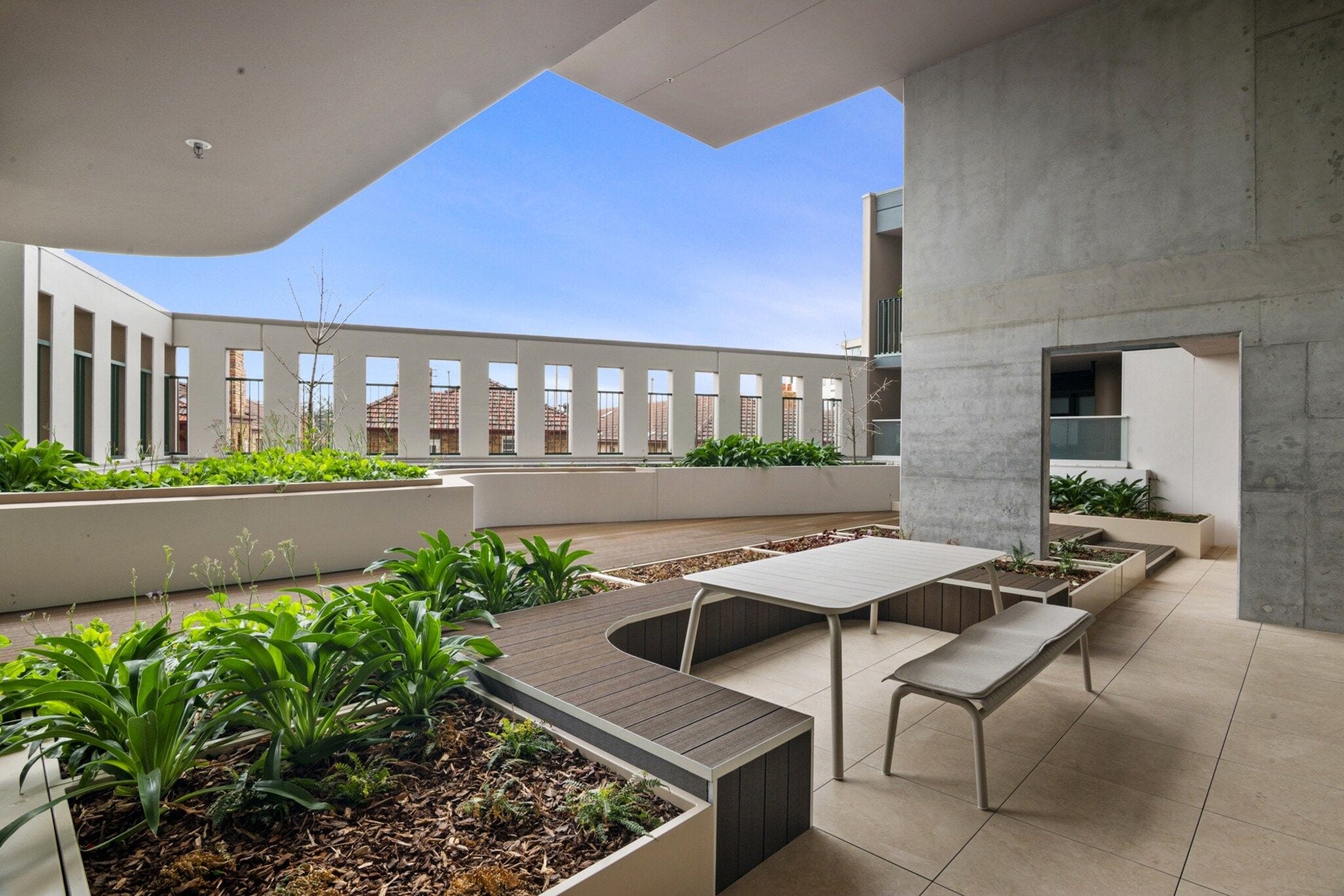 An outdoor patio with a modern design, featuring a seating area with benches and a table, surrounded by planters with greenery.