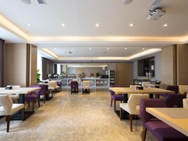 Un comedor moderno de hotel con varias mesas y sillas, una cocina al fondo e iluminación empotrada en el techo.