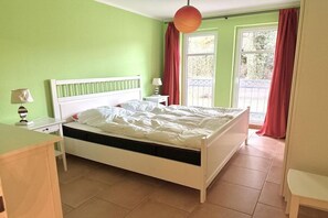 A bedroom with a white bed, a nightstand, a lamp, and a window with curtains. - Queen Augusta Dune Residence 2 (Ostseebad Binz)