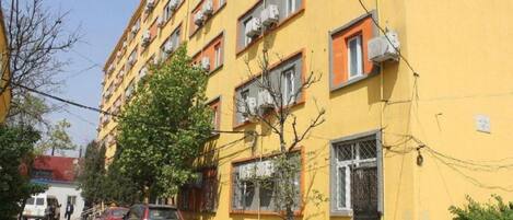A multi-story yellow building with air conditioning units, a tree in front, and parked cars along the street.