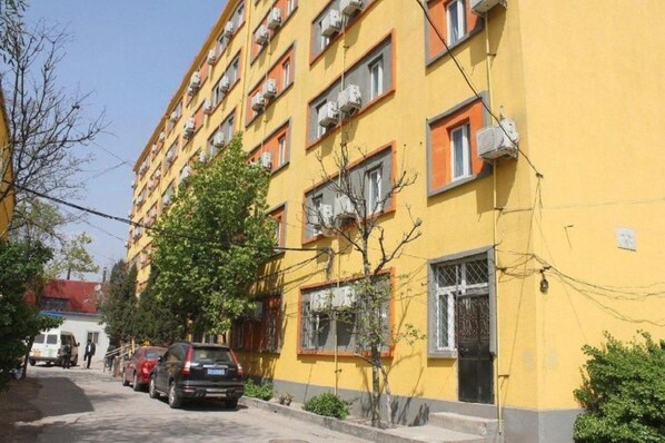A multi-story yellow building with air conditioning units, a tree in front, and parked cars along the street. - 7 Days Inn (Beijing)