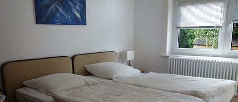Une chambre avec deux lits, un radiateur, une fenêtre avec des stores et un tableau accroché au mur.