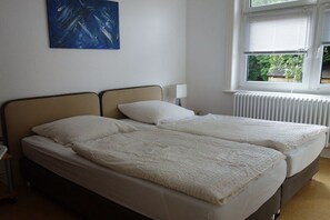 Ein Zimmer mit zwei Betten, einem Heizkörper, einem Fenster mit Jalousien und einem an der Wand hängenden Bild.