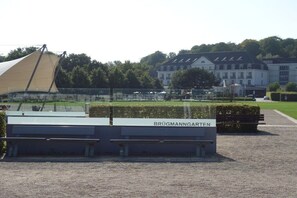 Ein Gebäude mit der Aufschrift 'BRÜGMANNGARTEN', links eine große Zelthalle, ein gepflegter Rasen und im Hintergrund ein klassisches Gebäude.