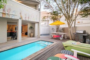 A poolside area with a wooden deck, a pool, and outdoor furniture including a table and chairs. - Green Point 4 Bed Family Home w Pool Happy House (Cape Town)
