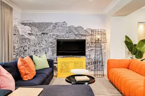 Een woonkamer met een zwart-wit behang, een flatscreen televisie, een geel kastje en een oranje bank.