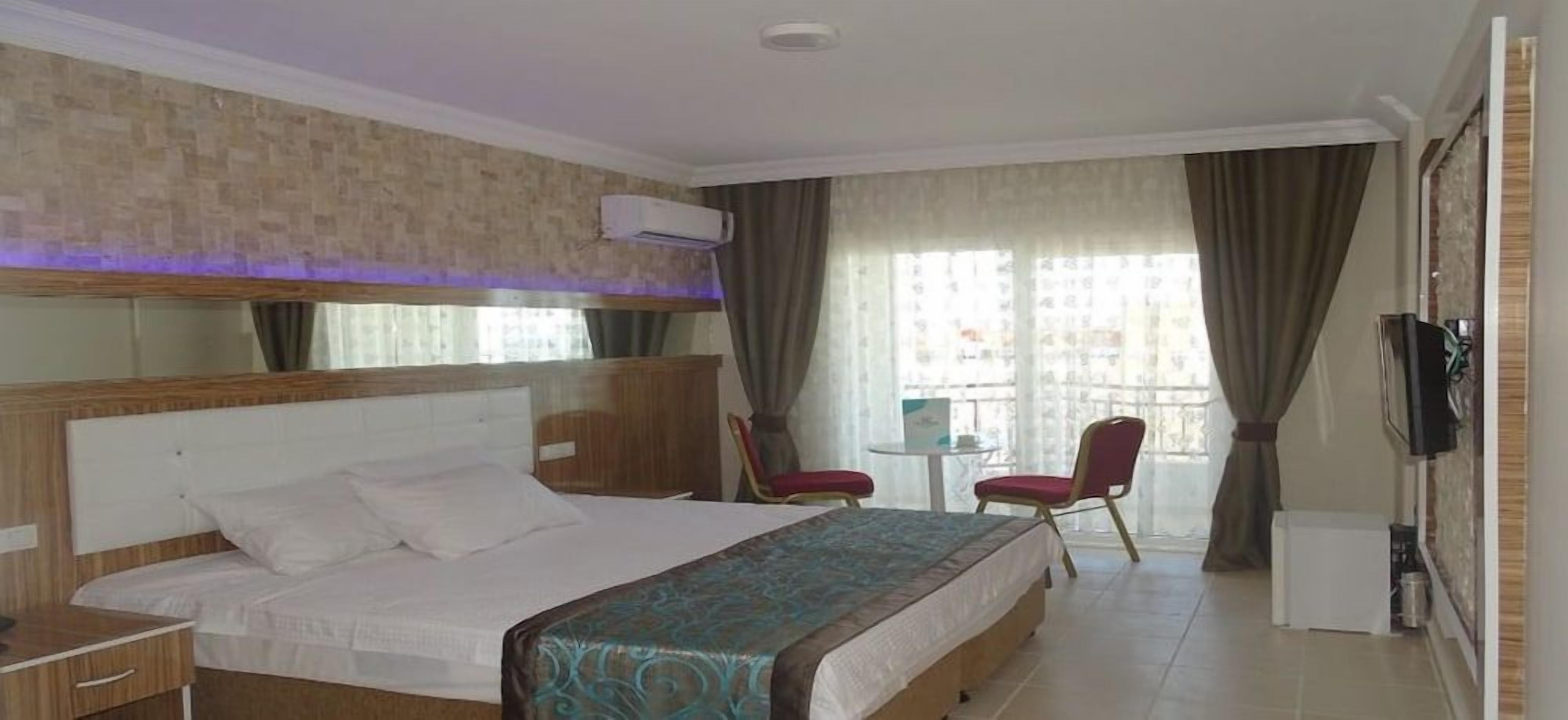 A hotel room with a large bed, a desk, a chair, a TV, and a balcony with curtains.