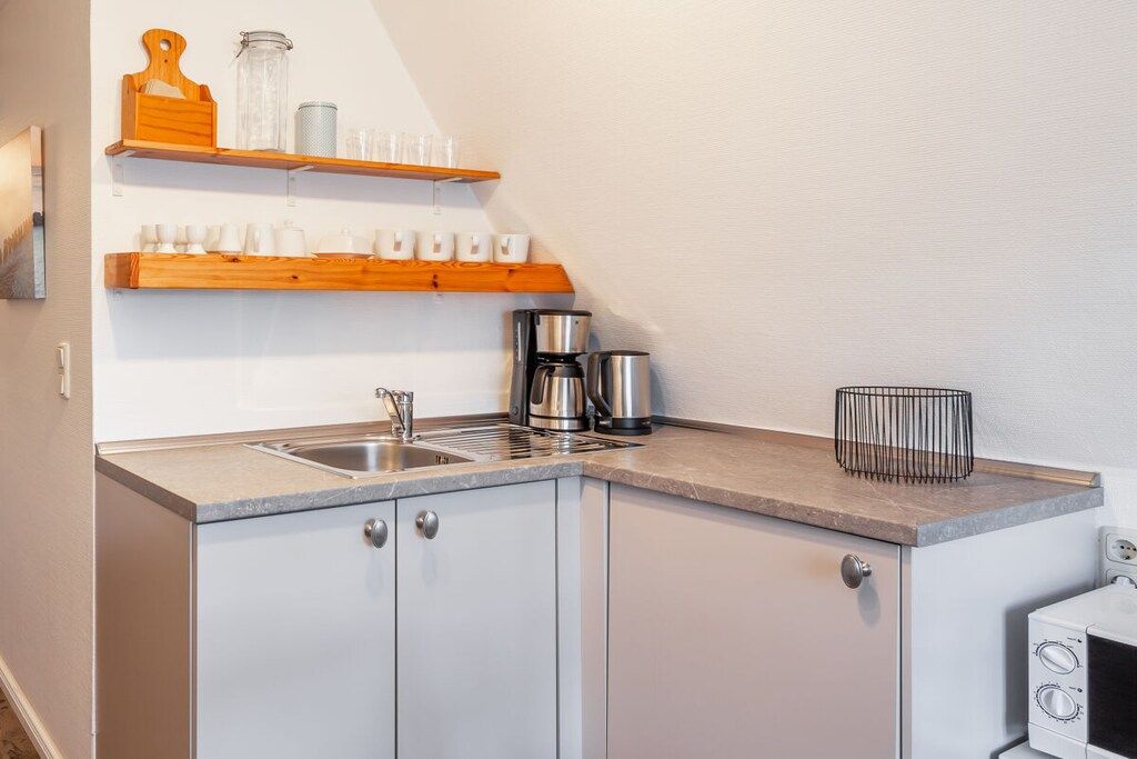 A compact kitchen with a sink, coffee maker, and a kettle. There are wooden shelves above the counter holding cups and a glass container.