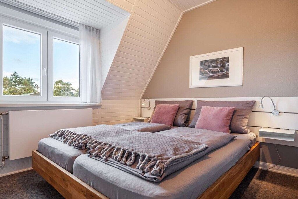 Une chambre avec un plafond en pente, un grand lit avec une tête de lit, une fenêtre avec des rideaux et un tableau encadré au mur.