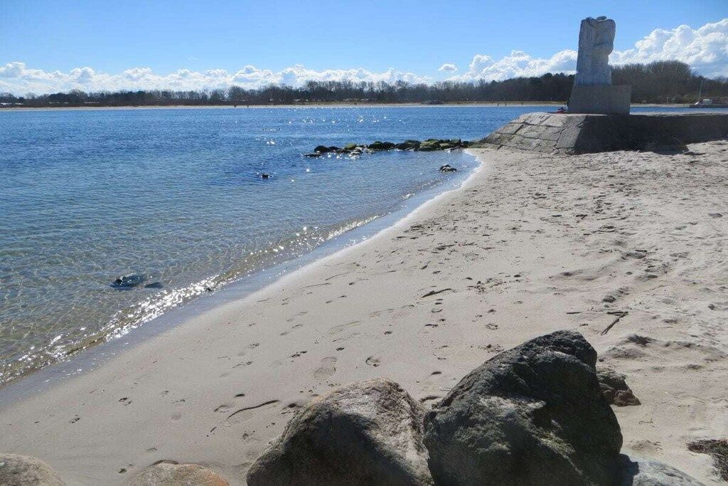A beach with clear water, a stone monument, and a distant tree line.