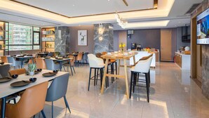 A modern dining area with multiple tables and chairs, a kitchen in the background, and a large window with a view of buildings. - Campanile (Shenzhen)