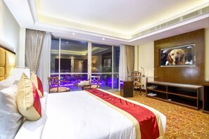 A hotel room with a large bed, a television, a seating area with a table and chairs, and a view through a glass door. - Grand Palace Rangpur (Rangpur)