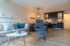 A modern living room with a sofa, armchair, and coffee table. A kitchen with dark cabinets and a dining area with a table and chairs is visible in the background. - Utholm 12 - Deichblick (St. Peter-Ording)