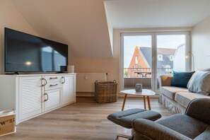 A modern living room with a flat-screen TV, a sofa set, a small table, and a view of a residential area through large windows. - Utholm 12 - Deichblick (St. Peter-Ording)