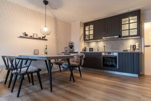 A modern kitchen with dark cabinetry, a built-in oven, and a dining area with a table and chairs. - Utholm 12 - Deichblick (St. Peter-Ording)
