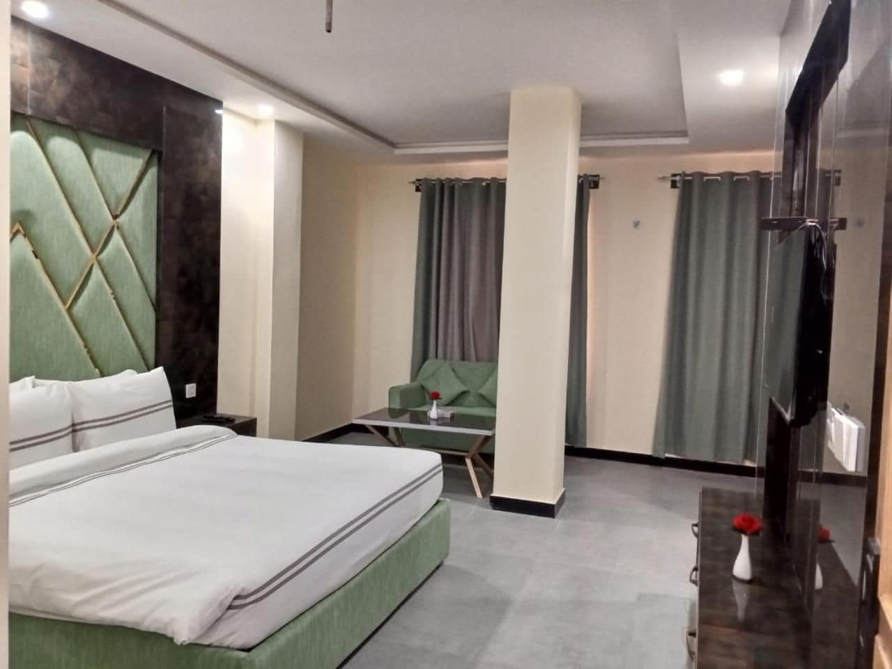 A hotel room with a bed, a green upholstered chair, a small table, and a mirror.