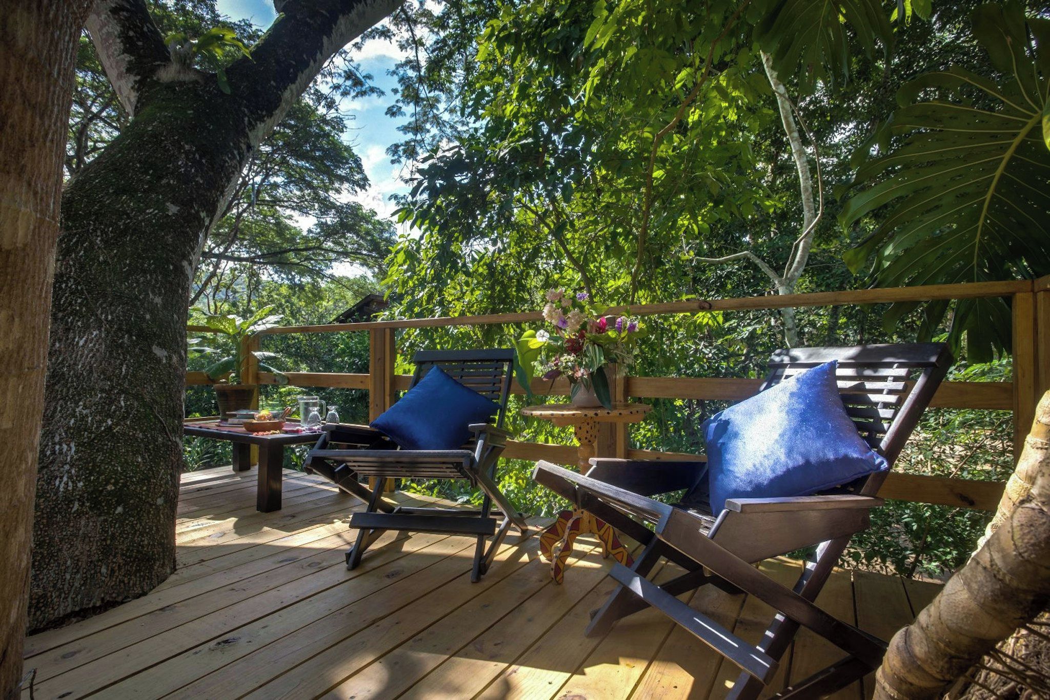 An outdoor deck with wooden furniture and a table, surrounded by lush greenery.