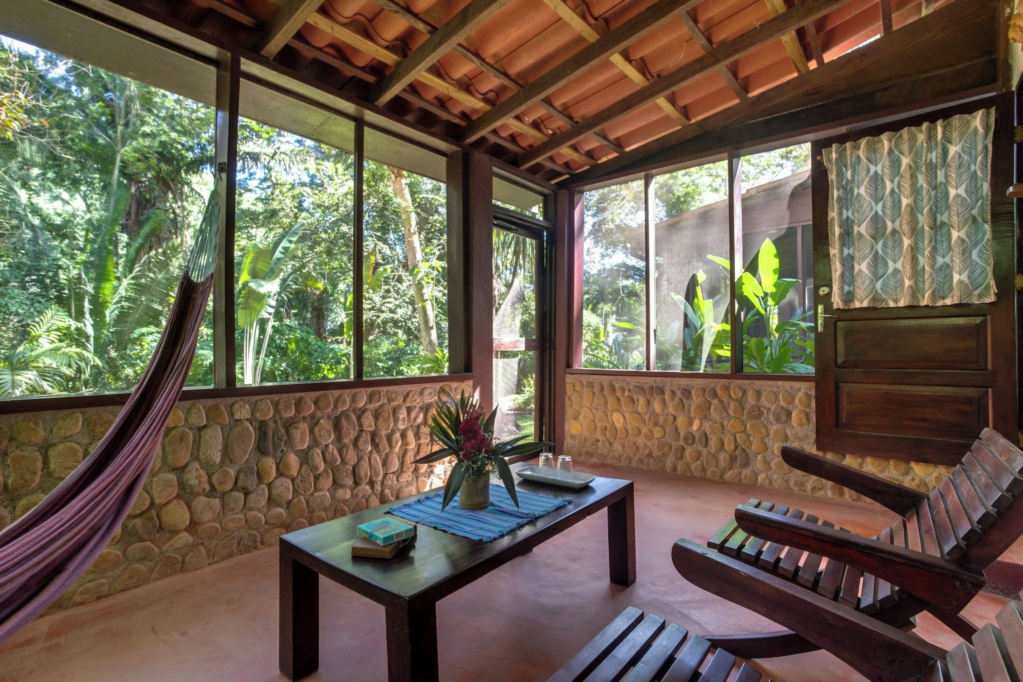 A room with a stone wall, a hammock, a wooden table with books and a plant, a wooden bench, and large windows with a view of greenery.