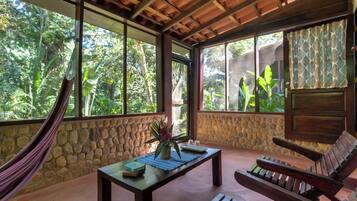 A room with a stone wall, a hammock, a wooden table with books and a plant, a wooden bench, and large windows with a view of greenery.