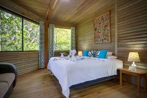 A wooden cabin bedroom with a bed, a nightstand, a lamp, and a window with curtains. - Sweet Songs Jungle Lodge (San Ignacio)