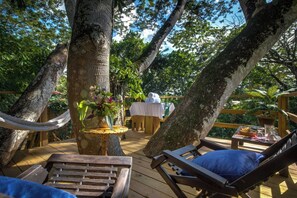 A wooden deck with lounge chairs and a table set for dining, surrounded by large trees. - Sweet Songs Jungle Lodge (San Ignacio)