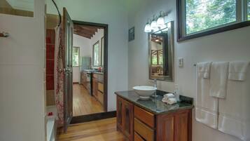 A bathroom with a wooden vanity, a sink, and a mirror with lights. There is a shower area with a curtain and a view of a dining area through an open door.