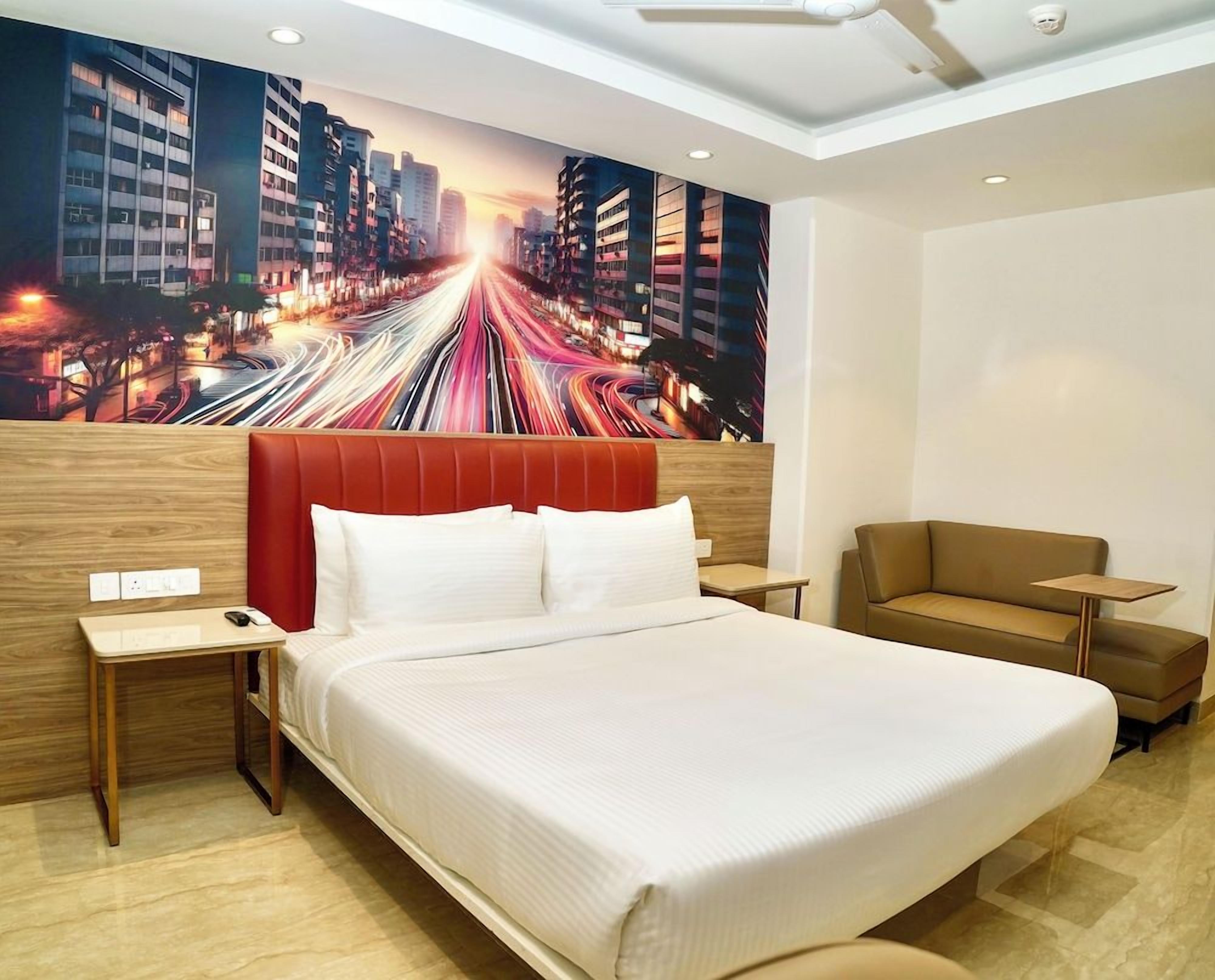 A hotel room with a large bed, a red headboard, a brown sofa, and a cityscape mural on the wall.