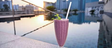 A glass of a purple beverage with a straw and mint garnish on a balcony with a pool and city buildings in the background.