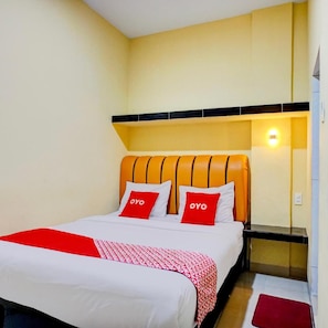 Habitación de hotel con cama, dos almohadas con la palabra 'OYO', una manta roja y blanca estampada y una mesita de noche con lámpara.