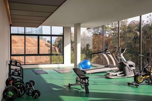 Un gimnasio con equipo de ejercicio, una banca y un amplio ventanal con vista a una colina y árboles.