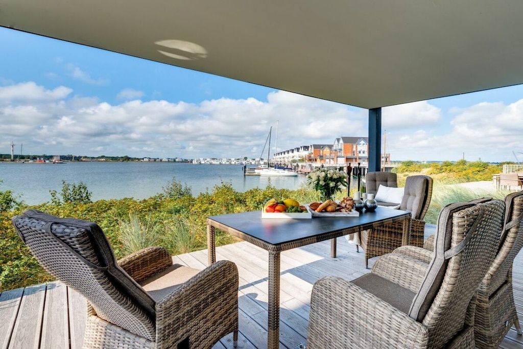 An outdoor deck with wicker furniture, a table with fruits and a vase, and a view of the water and buildings in the distance.
