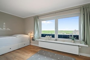 Ein Schlafzimmer mit einem Bett, einem Fenster mit Vorhängen, einem Heizkörper und Blick ins Freie.