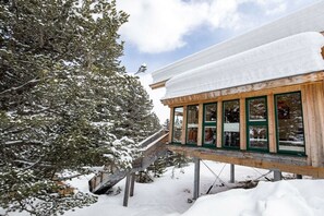 Un edificio de madera con amplios ventanales, una terraza de madera y un techo cubierto de nieve.