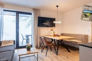 A modern living room with a sofa, a dining table with chairs, a TV mounted on the wall, and a large window with curtains. - Holiday apartment & infinity pool (St. Martin am Tennengebirge)